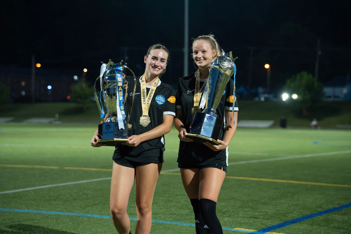 Two soccer players holding trophies and wearing medals on the field